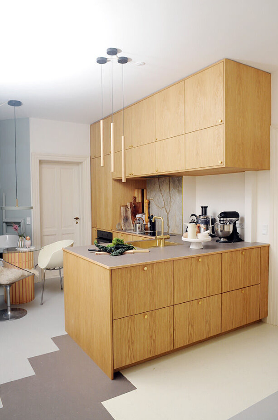 Køkkenskabene er lavet af Ikea-indmad beklædt med snedkerbyggede låger i egetræsfinér, hvor årerne løber gennemgående fra gulv til loft. Bordpladen er beklædt med linoleum fra Forbo, den er slidstærk og giver et blødt og roligt udtryk i køkkenet. Messingvasken er fra Lavabo og købt hos billigvvs.dk, hvor man også kan købe messingarmaturet fra Damixa. De smukke lange egetræsrør over bordpladen, er pendler fra Umage Design.