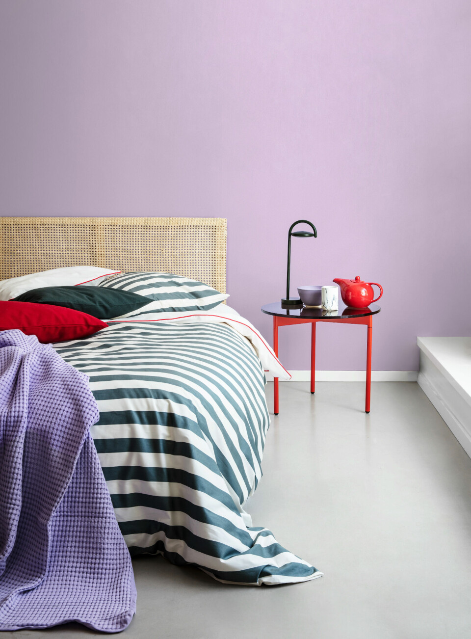 Stribet dynebetræk, Hay, 449 kr. Stribet pudebetræk, Hay, 99 kr.Hvidt sengelinned med rødt kantbånd, Ikea, 179 kr. Lilla sengetæppe, Ikea, 229 kr. Grøn pude, H&M home, 39 kr. Rød pude, H&M home, 39 kr. Væggen er malet med farven Purple Drogon fra Dyrup. Lille bord med glasplade, Warm Nordic, 1.499 kr. Bordlampe, Hay, 1.199 kr.