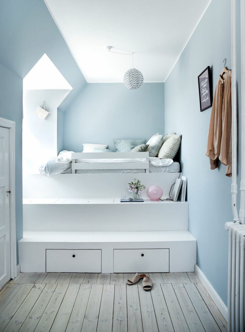 For at man kan ligge i sengen og kigge ud af vinduet, er sengen hævet op på flere niveauer af trappetrin med låger og lange, dybe skuffer. Væggene er malet lyseblå i farven Parma Grey fra Farrow and Ball. Grebene er fra Københavns Møbelsnedkeri og loftslampen er fra Dora.