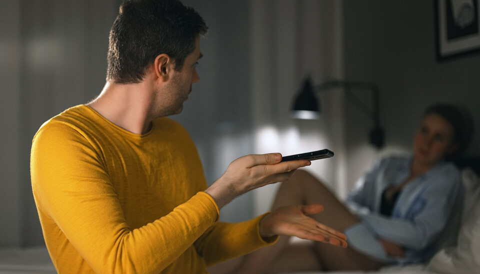 En mand sidder med sin telefon i hånden, i en seng, mens han diskuterer med sin kone.