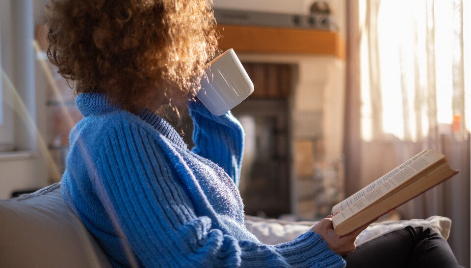 Kvinde drikker af en kop mens hun læser i en bog i sin sofa.
