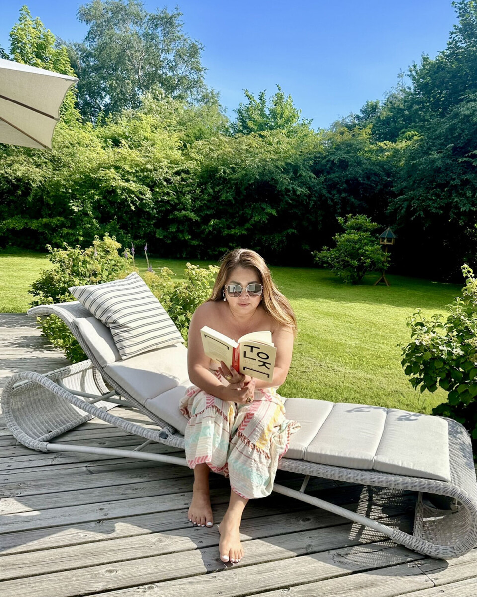 Ann Lind Andersen sidder udenfor i solen, og hun læser i en bog.