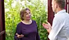 En ældre kvinde med en lilla trøje på, står og diskuterer med en yngre herre. Han har en skjorte på, som er hvid. I baggrunden kan man se et grønt areal.