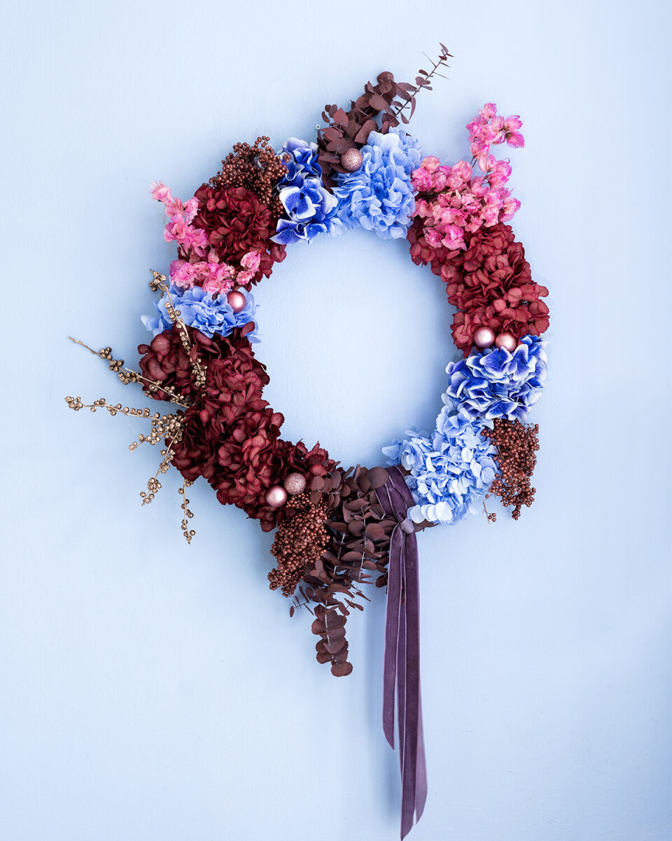 I kransen fra Holly Flower Studio er der brugt både friske og kunstige hortensia, spray-malede peberkorn, farvet eucalyptus, malede ilexgrene, bourgonvilla og et par julekugler. Bemærk velourbåndet, der hænger ned fra kransen som en ekstra dekorativ detalje.