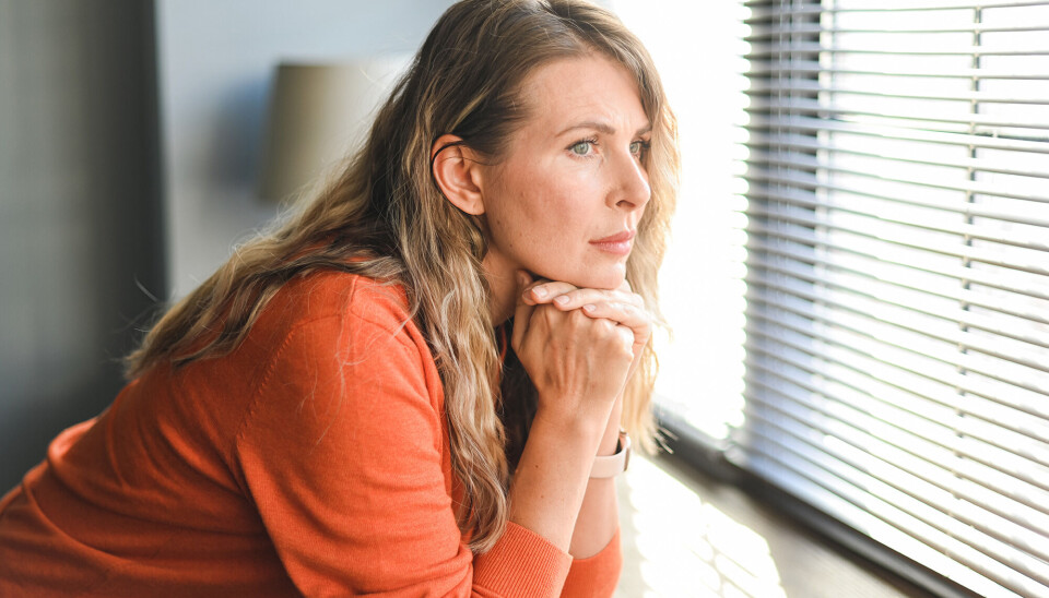 En kvinde, som har en orange trøje på. Hun støtter sine albuer på vindueskarmen, og ser ud af vinduet.