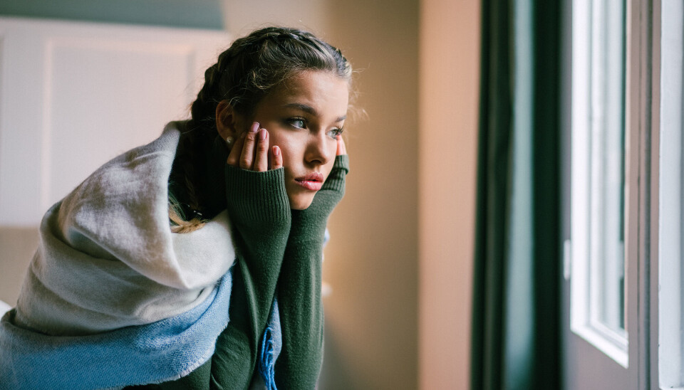 En ung kvinde sidder på en seng. Hun har et tæppe viklet rundt om sig, og hun ser fortvivlet ud. Hun kigger ud af vinduet, mens hun læner sit hoved i hænderne.