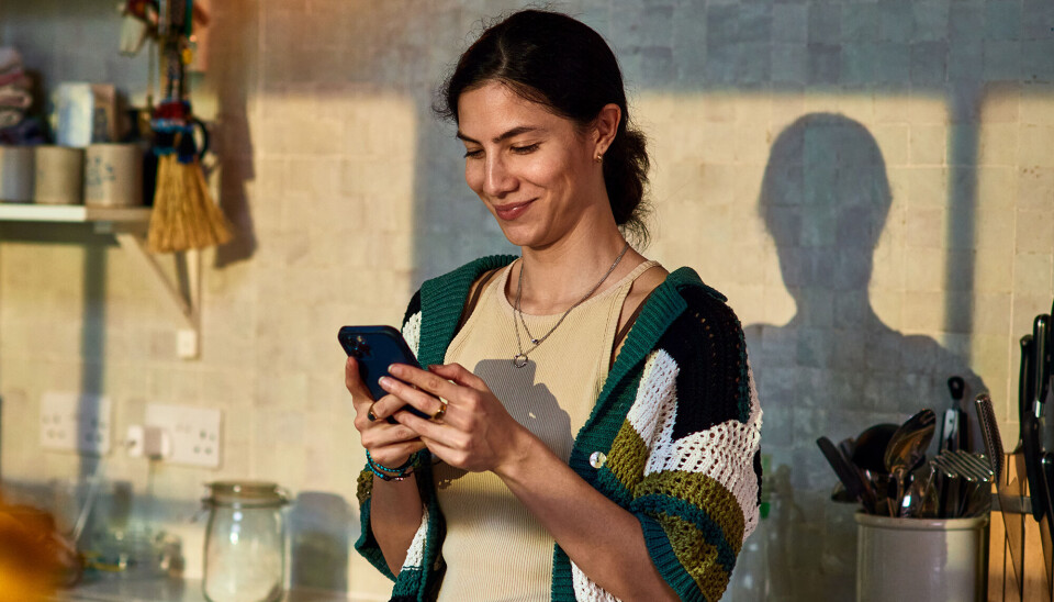 En kvinde, som står i sit køkken. Hun ser ned på sin telefon, mens hun smiler. Hun har en lys top på, og en cardigan i forskellige farver over.