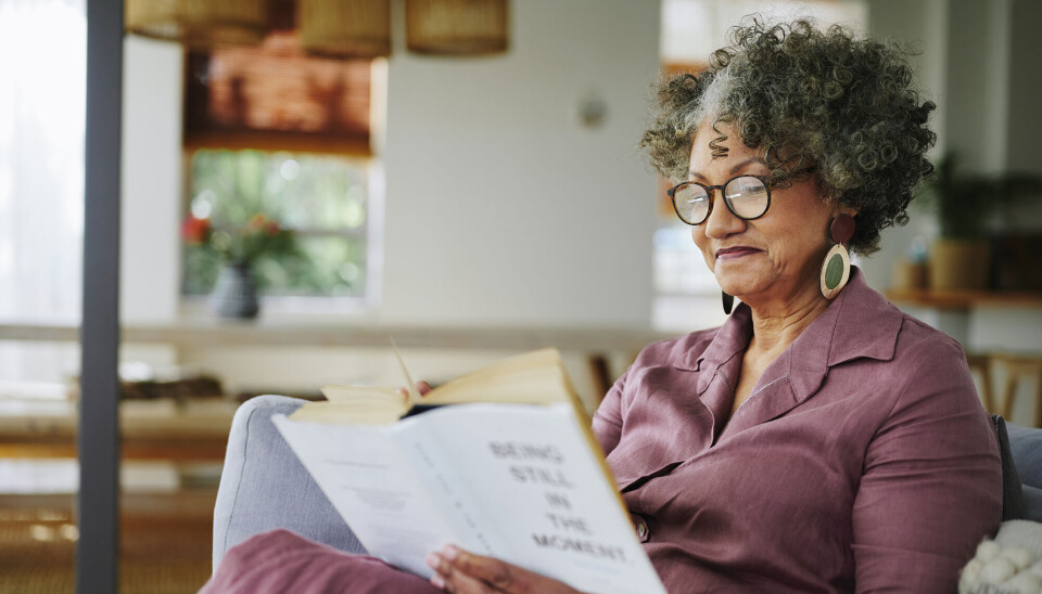 En kvinde sidder i sin sofa og læser en bog. Hun har en rosa kjole på, briller og har mørkt, krøllet hår.