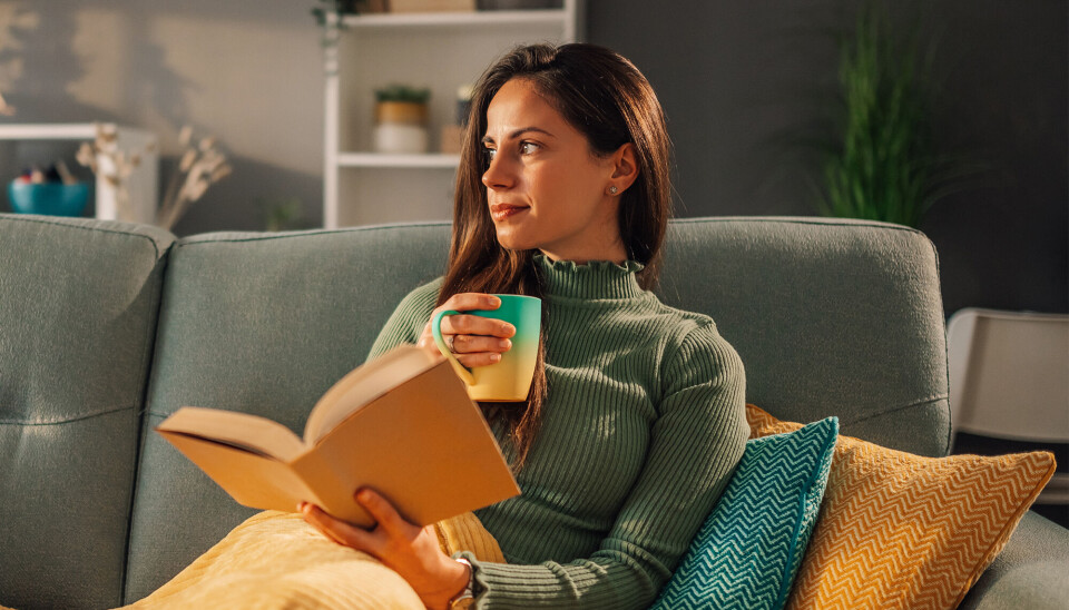 Portræt af en ung kvinde, som sidder i en behagelig sofa mens hun læser en bog og drikker en kop te eller kaffe. Hun har et tæppe over sig.