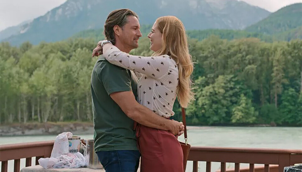 Stilbillede fra serien 'Virgin River'. Her ses hovedpersonen med en mand, mens de holder om hinanden. De står ude ved en flod, på en restaurant og der er livlig natur i baggrunden med bjerge ude i horisonten.