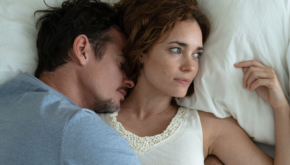 En mand og en kvinde ligger i en seng. Kvinden ser væk, og manden ligger helt tæt på hende. Hun ser utilpas ud.