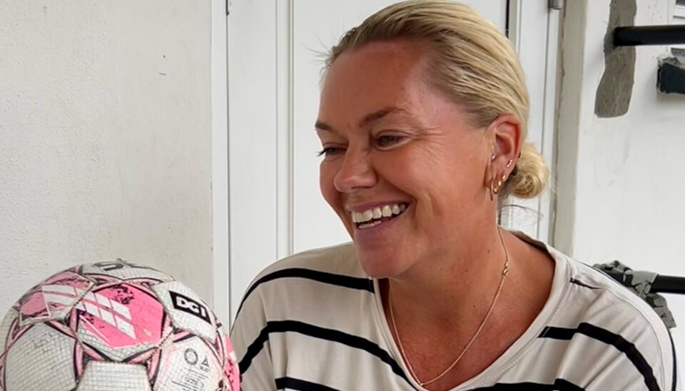 Katrine Memborg sidder med en fodbold, som er hvid og lyserød. Hun har en hvid bluse på, med sorte striber. Hun har sit lyse hår i en lav knold, og hun smiler.