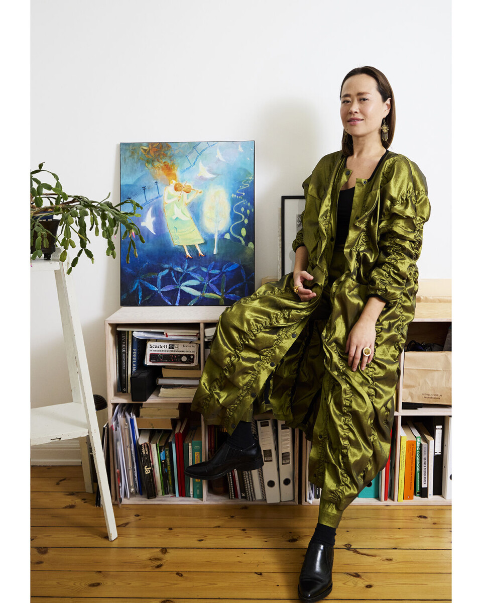 Billede af Ida Kudo. Hun sidder på en kommode, og har et sæt på, som er grønt. I kommoden ses bøger og mapper, og på kommoden står et maleri.