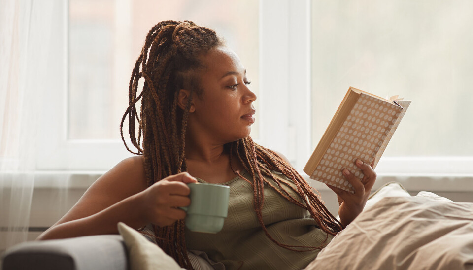 En ung kvinde ligger i sin sofa og læser i en bog. Hun har mørkt hår, med mange små fletninger. Hun holder et lyseblåt kaffekrus.