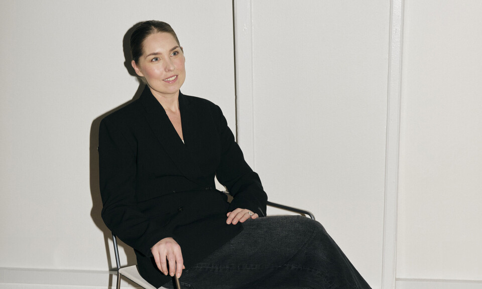 Billede af Andrea Hess. Hun sidder på en stol, har et sort jakkesæt på, og væggen er helt hvid.