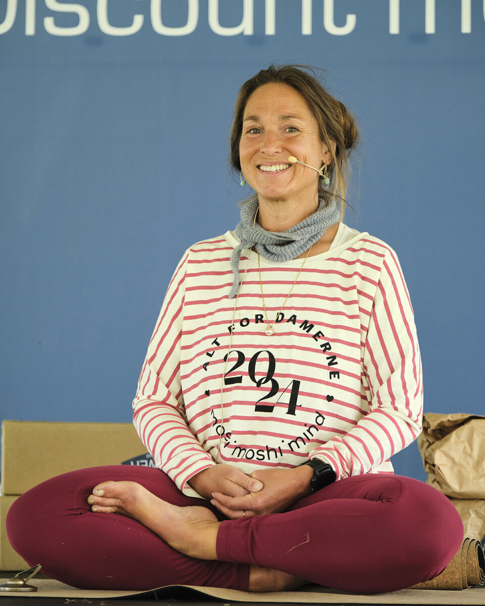 Billede af Kristine Marie Rost, som underviser i Yin yoga. Billedet er taget til sidste års ALT for damernes yoga-event. Hun har en hvid/rødstribet trøje på, hvor der er trykt 'ALT for damerne, 2024' i sort skrift på trøjen. Hun sidder i skrædderstilling, og har et gråt Sophie-scarf rundt om halsen.