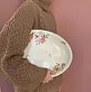 Fra siden ses en kvinde iført en stor, brun sweater. Man kan kun se kroppen, ikke hovedet. Hun har et stort porcelænsfad med blomster på under armen.