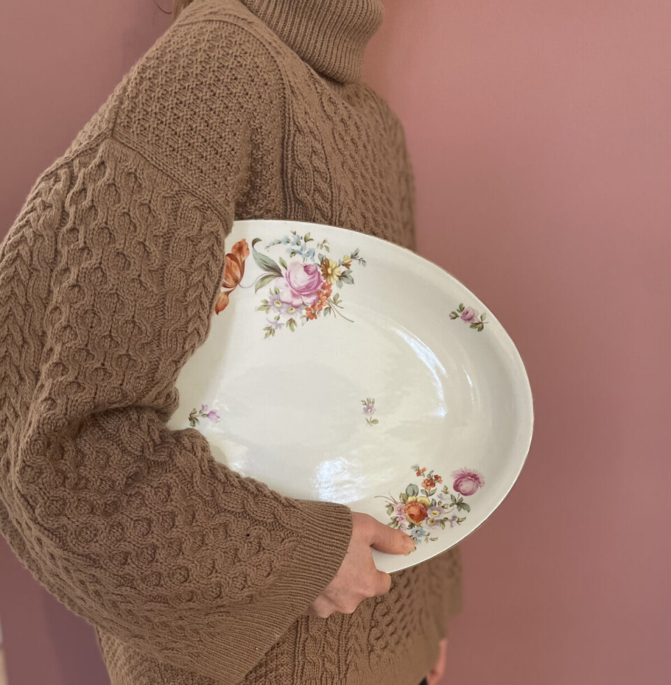 Fra siden ses en kvinde iført en stor, brun sweater. Man kan kun se kroppen, ikke hovedet. Hun har et stort porcelænsfad med blomster på under armen.
