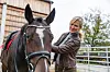 An adult blonde female horse trainer looking at her horse and caressing it. They are standing outside in front of the stables. They are surrounded by beautiful nature and a fence. The woman is wearing fall clothes. She looks focused. The weather is cloudy.