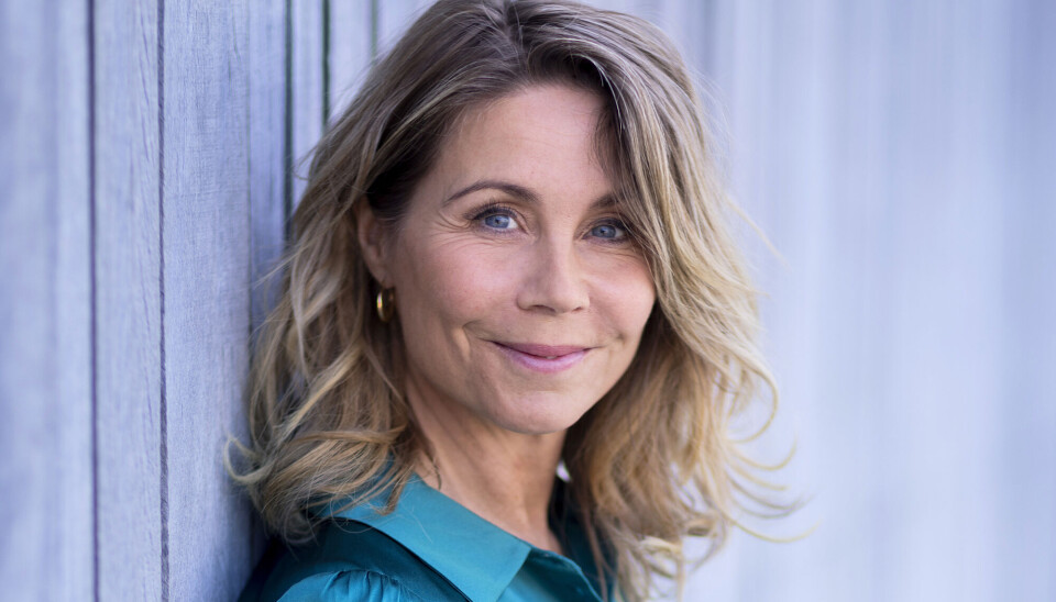 Portræt af Anne Louise Hassing. Hun står op ad en trævæg, som er blålig. Hun har løst, lyst hår og smiler direkte ind i kameraet. Hun har en blå skjorte på i satin.