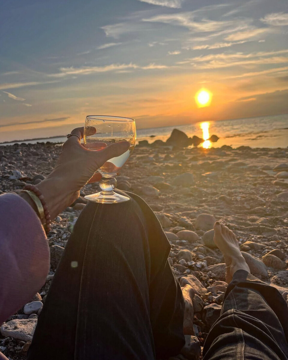 Anne Louise Hassing har taget et billede af, at hun ligger på stranden, og ser på solnedgangen. Hun holder et glas vin med sin venstre hånd, og man ser kun hendes arme og ben. Det er en stenstrand.