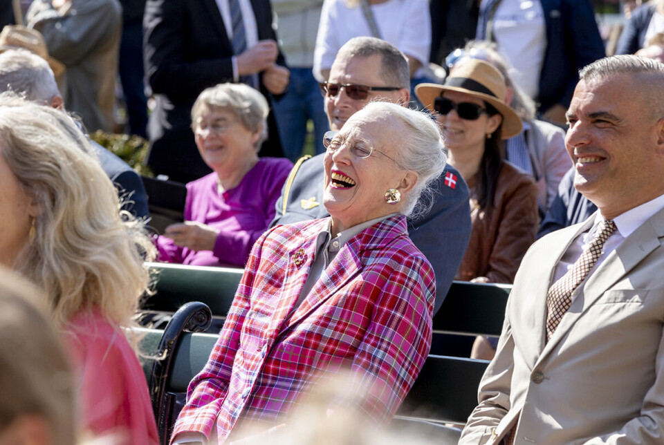Dronning Margrethe i ternet blazer i rød og lyserød til Pantomimeteatrets 150-års jubilæum og afsløring af nyt påfugle-fortæppe. Dronningen sidder på en tilskuerbænk og griner med hovedet lænet bagover.