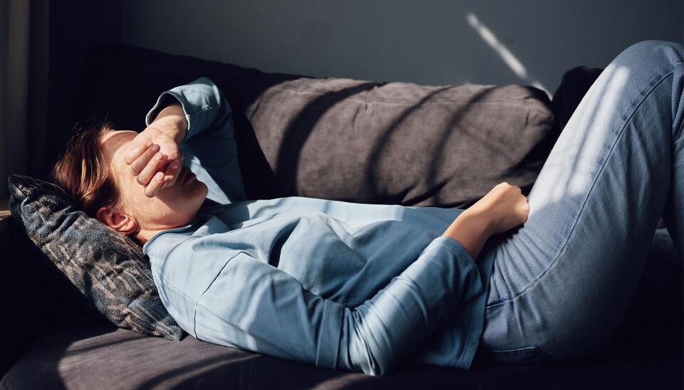 En kvinde i 30'erne ligger på en sofa. Hun har en blå trøje og blå bukser på. Hun holder sin ene hånd over sit hoved. Sofaen er mørkegrå.