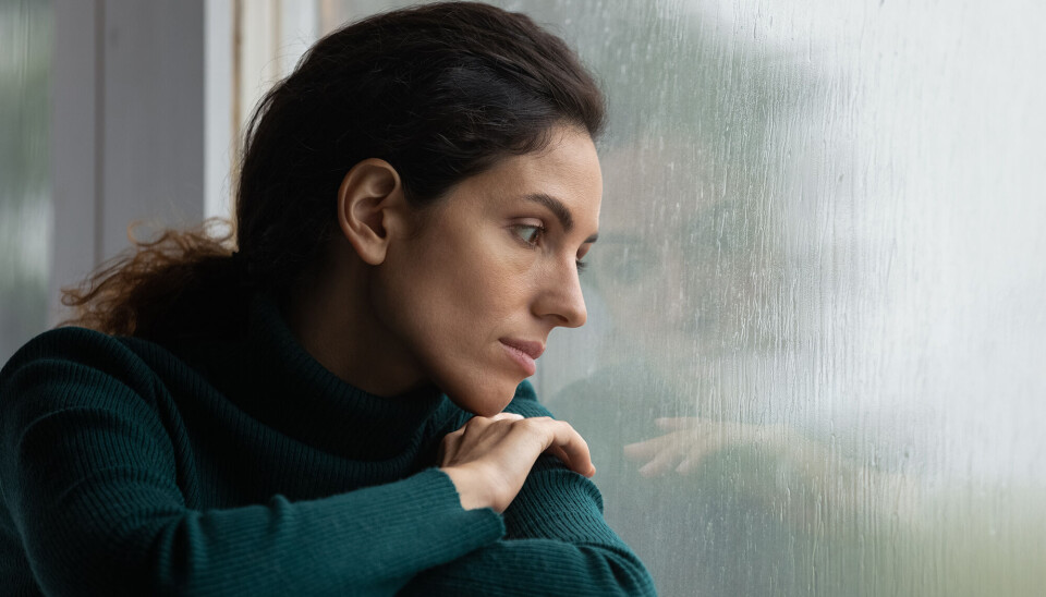 En kvinde i 30'erne sidder og kigger ud af vinduet. Hun har mørkt, langt hår, som er sat op i en lav hestehale. Hun har en mørkegrøn langærmet trøje på. Hun ser trist ud.