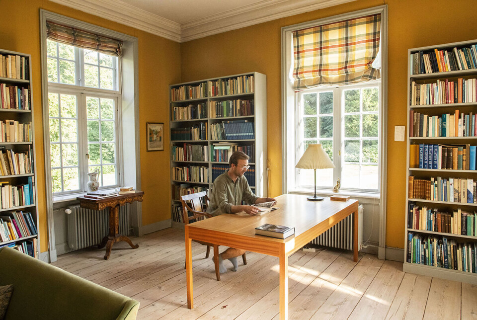August Hage sidder bag et skrivebord i et stort, lyst rum med bogreoler langs væggene.