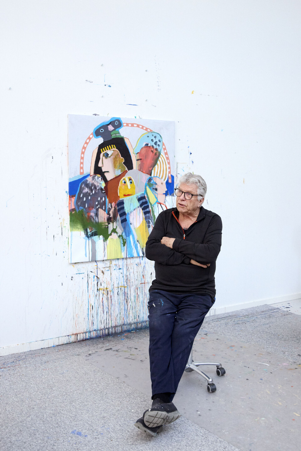 Foto af Leif Sylvester. Han sidder i sit atelier, og bag ham hænger et maleri på væggen i mange forskellige farver.