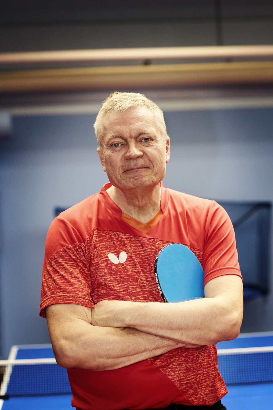 Træner Lars Bo iført rød trøje med armene over kors. I hånden har han et lyseblåt bordtennisbat.