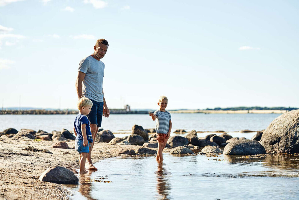 En mand med to smø børn står i vandkanten. Omkring dem er der store sten.