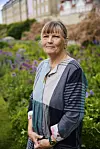Forfatter Anna Grue i en mønstret kjole foran et blomsterbed.