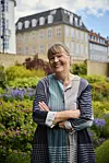 Forfatter Anna Grue i en mønstret kjole foran et blomsterbed.