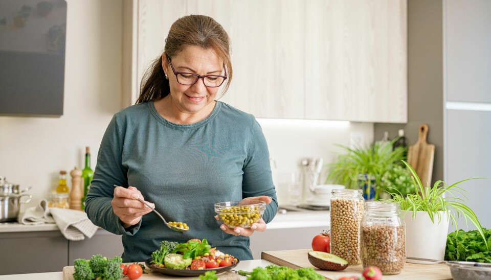 En kvinde på omkring 50. Hun står i et køkken, og er ved at lave mad. På billedet er der broccoli, kikærter, avokado og tomater.