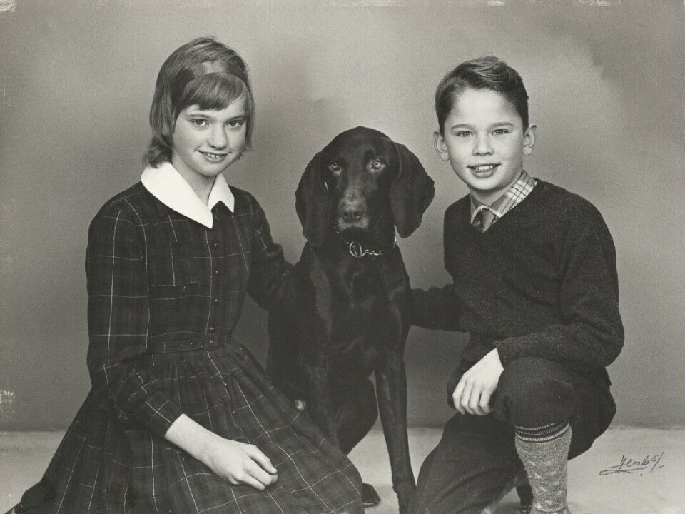 Pia sidder på gulvet iklædt en ternet kjole og med en hund i midten, mens hendes lillebror er sidder på hug til højre. Han er iklædt en ternet skjorte. De smiler ind i kameraet.