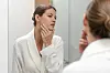 Kvinde i hvid morgenkåbe tjekker huden på sin kind foran badeværelsesspejl.