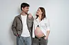 Gravid kvinde med åben skjorte står smilende ved siden af sin partner mod lys baggrund
