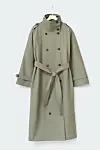 Olivengrøn lang trenchcoat med bælte hænger på en bøjle mod lys baggrund