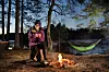 Linda ved lejrbål i skov med hængekøje og sø i baggrunden i skumringen.