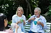 To ældre personer drikker kaffe på en bænk i en grøn have og taler med en ven.