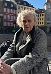 Ældre kvinde sidder udendørs i en grå jakke foran farvede huse i København.