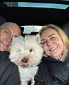 Par i bil tager selfie med lille hvid hund mellem sig på forsædet