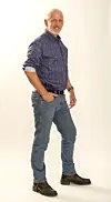 Ældre mand i skjorte, jeans og støvler poserer stående mod lys baggrund.