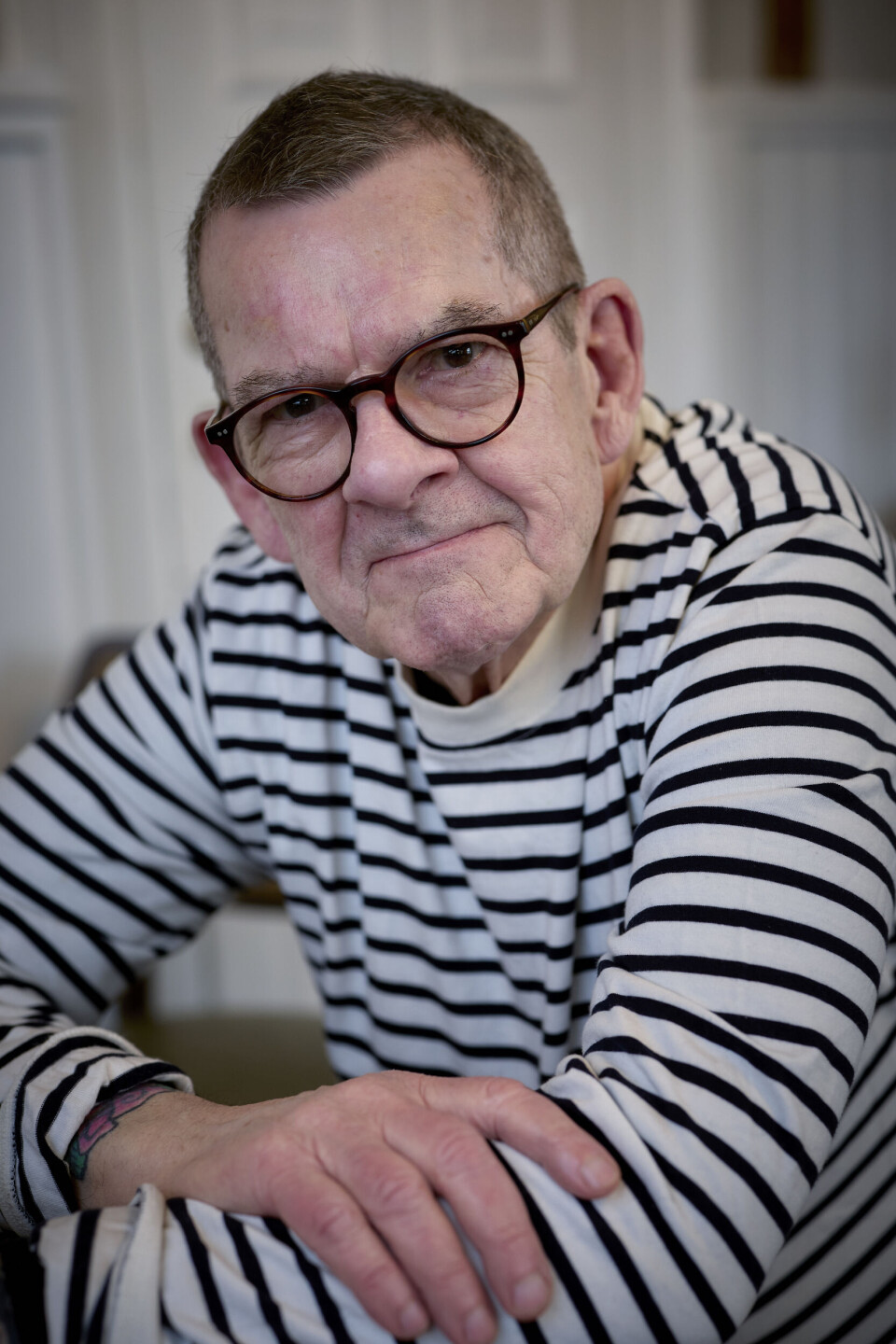 Billede 6 - Ældre mand med briller i sort-hvid stribet trøje sidder indendørs og ser mod kameraet