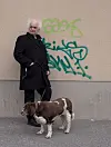 Person i mörk kappa står med hund i koppel framför en graffitimålad vägg.
