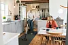 A man vacuuming the kitchen while his wife drinks coffee.