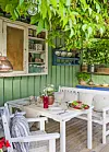 Romantisk siddeplads under tag:
Kurvemøbler med bløde former samt hynder og puder indbyder til afslappende eftermiddage på siddepladsen ved huset. Træterrassen i grå passer flot til møblerne og de rosa detaljer. I det gamle træ hænger rosa pelargonier i hængekrukker, og på bænken står en lanterne, som kan tændes om aftenen.