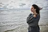 Kvinde i stor grå sweater står alene på stranden og ser ud over havet