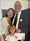 Tre personer står tæt sammen indendørs, mens barnet holder et farverigt kort frem.